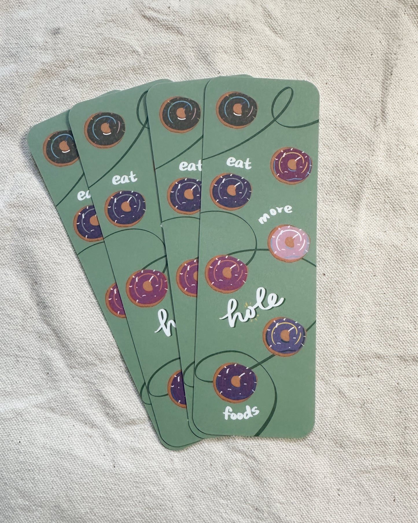 A handmade bookmark with subtle pride colored donuts placed on a sage green background. The donuts represented are the progressive pride flag, pansexual, asexual, transgender, lesbian, nonbinary, bisexual flag colors. There is writing that says "eat more hole foods" as a play on "eat whole foods" and "hole" for donuts as a food with holes